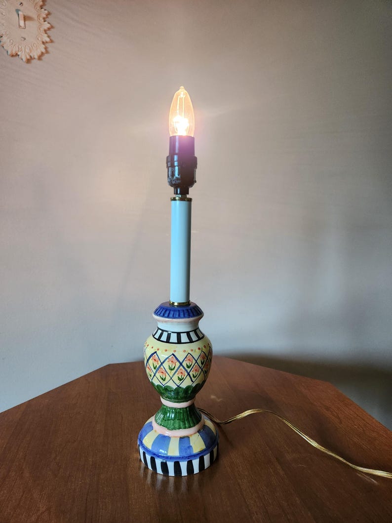 May include: A decorative table lamp with a lit bulb. The lamp has a light blue stem and a colorful base with yellow, green, blue, and black patterns. The lamp is on a wooden surface.