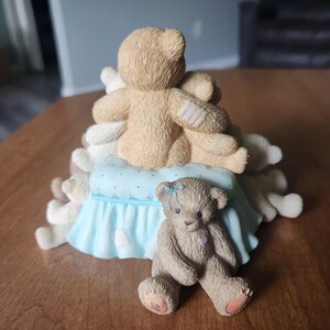 Puede incluir: Una figura decorativa con tres osos de peluche. Dos osos se abrazan sobre una almohada azul claro con una falda con volantes. Un tercer oso est&aacute; sentado al frente, con un lazo azul y un coraz&oacute;n rosa.