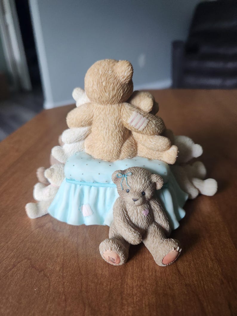 Puede incluir: Una figura decorativa con tres osos de peluche. Dos osos se abrazan sobre una almohada azul claro con una falda con volantes. Un tercer oso est&aacute; sentado al frente, con un lazo azul y un coraz&oacute;n rosa.