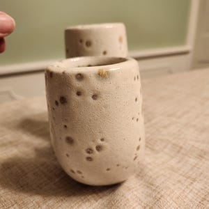 May include: Two handmade ceramic cups with a speckled, textured surface. The cups are off-white with brown spots and a rounded shape. The cups are likely for drinking beverages.
