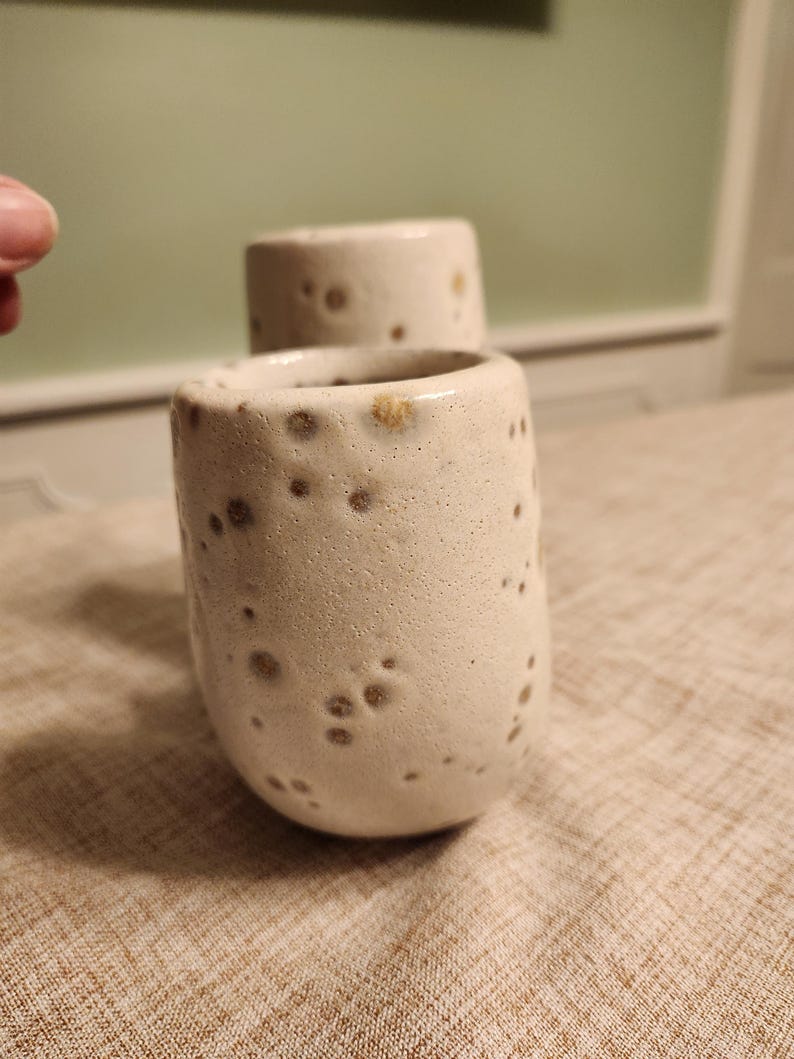 May include: Two handmade ceramic cups with a speckled, textured surface. The cups are off-white with brown spots and a rounded shape. The cups are likely for drinking beverages.