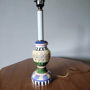 May include: A decorative table lamp with a colorful, patterned ceramic base. The base features a green, yellow, blue, and black design. The lamp has a white central column and a black lamp holder. The lamp is on a wooden table.