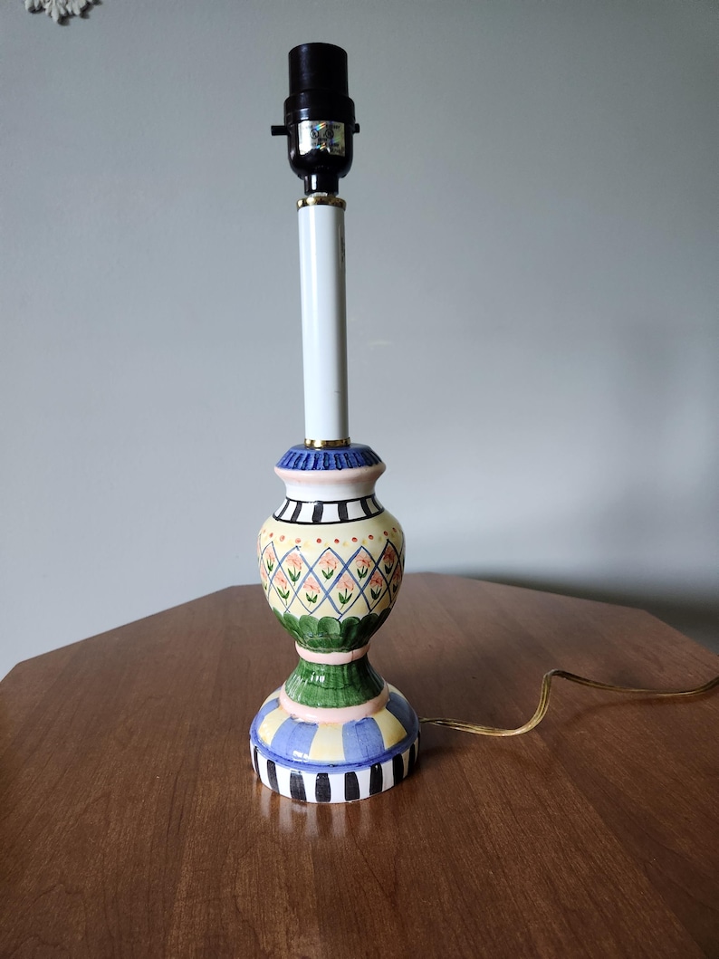 May include: A decorative table lamp with a colorful, patterned ceramic base. The base features a green, yellow, blue, and black design. The lamp has a white central column and a black lamp holder. The lamp is on a wooden table.