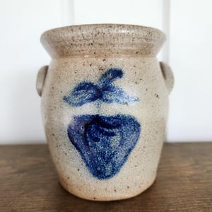 May include: A speckled beige stoneware crock featuring a hand-painted blue strawberry design on the front. The vessel has a wide, flared rim and two small side handles, with a textured, earthy finish throughout the exterior surface.