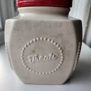May include: A beige ceramic jar with a red lid. The jar has a hexagonal shape with a crackle finish and the word "Treats" in a dotted circle. The jar is designed for storing treats.