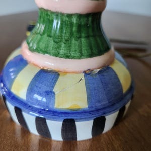 May include: A colorful ceramic decorative vase with a green top, a pink band, and a base with blue and yellow stripes. The bottom of the vase has black and white vertical stripes. The vase is on a brown surface.