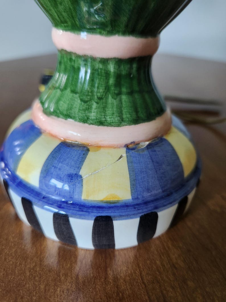 May include: A colorful ceramic decorative vase with a green top, a pink band, and a base with blue and yellow stripes. The bottom of the vase has black and white vertical stripes. The vase is on a brown surface.