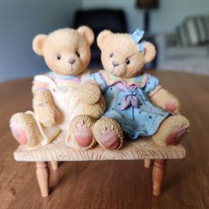 May include: A decorative figurine featuring two teddy bears seated on a small wooden bench. One bear wears a light blue dress with a bow, the other a pastel striped top. The bears have tan fur and pink paw pads.