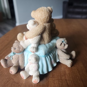 Puede incluir: Una figura decorativa con varios osos de peluche. Los osos son de color marr&oacute;n y blanco, algunos con lazos azules o rosas. Est&aacute;n colocados sobre una base azul claro con un borde con volantes.