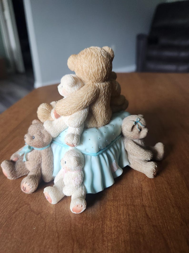 Puede incluir: Una figura decorativa con varios osos de peluche. Los osos son de color marr&oacute;n y blanco, algunos con lazos azules o rosas. Est&aacute;n colocados sobre una base azul claro con un borde con volantes.