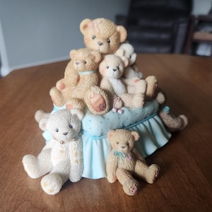 Puede incluir: Figura decorativa con un grupo de ositos de peluche en varias poses y tama&ntilde;os. Los osos son principalmente de color beige y crema, algunos con peque&ntilde;os lazos. Est&aacute;n colocados sobre una base azul claro con una falda con volantes.