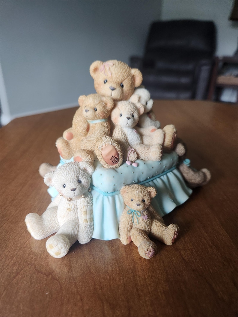 Puede incluir: Figura decorativa con un grupo de ositos de peluche en varias poses y tama&ntilde;os. Los osos son principalmente de color beige y crema, algunos con peque&ntilde;os lazos. Est&aacute;n colocados sobre una base azul claro con una falda con volantes.