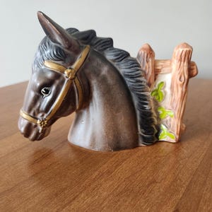 Könnte beinhalten: Ein Keramik-Pferdekopf-Pflanzgefäß in Braun und Schwarz. Das Pferd hat ein goldenes Zaumzeug und eine schwarze Mähne. Ein Holzzaun mit grünen Blättern befindet sich hinter dem Pferdekopf.