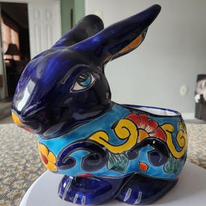 May include: A ceramic rabbit planter with a vibrant blue glaze. The planter features a detailed floral design in yellow, red, and green. The rabbit has long ears and a detailed face, perfect for adding a touch of whimsy to any space.