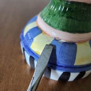 May include: A colorful ceramic vase with a striped and patterned design. The vase features a blue, yellow, green, pink, and black color scheme. A metal tool is placed near the vase.