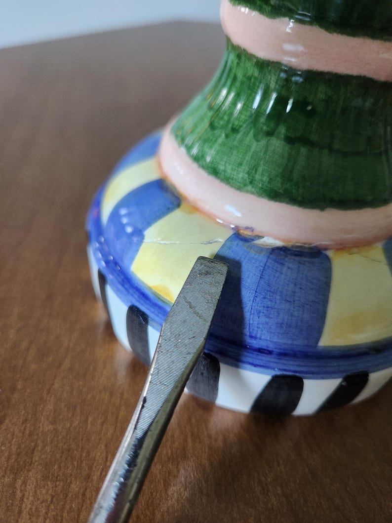May include: A colorful ceramic vase with a striped and patterned design. The vase features a blue, yellow, green, pink, and black color scheme. A metal tool is placed near the vase.