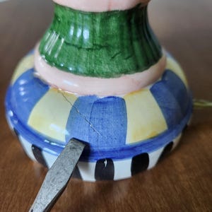May include: A colorful ceramic decorative item with a green, pink, blue, yellow, and black striped design. The item has a crack and a metal tool is placed near it.