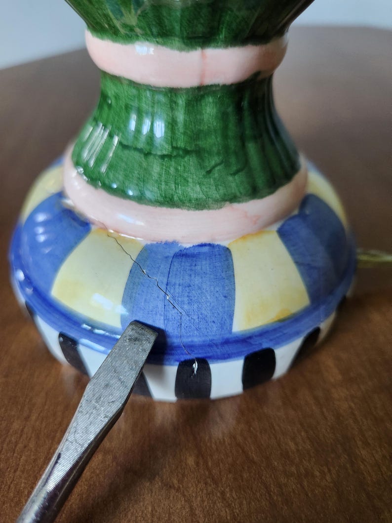 May include: A colorful ceramic decorative item with a green, pink, blue, yellow, and black striped design. The item has a crack and a metal tool is placed near it.