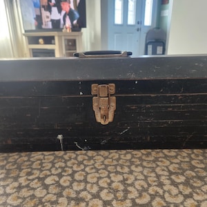 May include: A vintage black metal toolbox with a hinged lid and a metal clasp. The toolbox has a handle on top and shows signs of wear, with some rust and paint chipping. The toolbox is rectangular and appears to be used for storing tools.