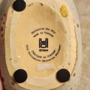 May include: Close-up of the bottom of a speckled, light green ceramic vase. The base is beige with three black circular pads. Text reads "Decorative Use Only Made in Vietnam" and "UG Home".