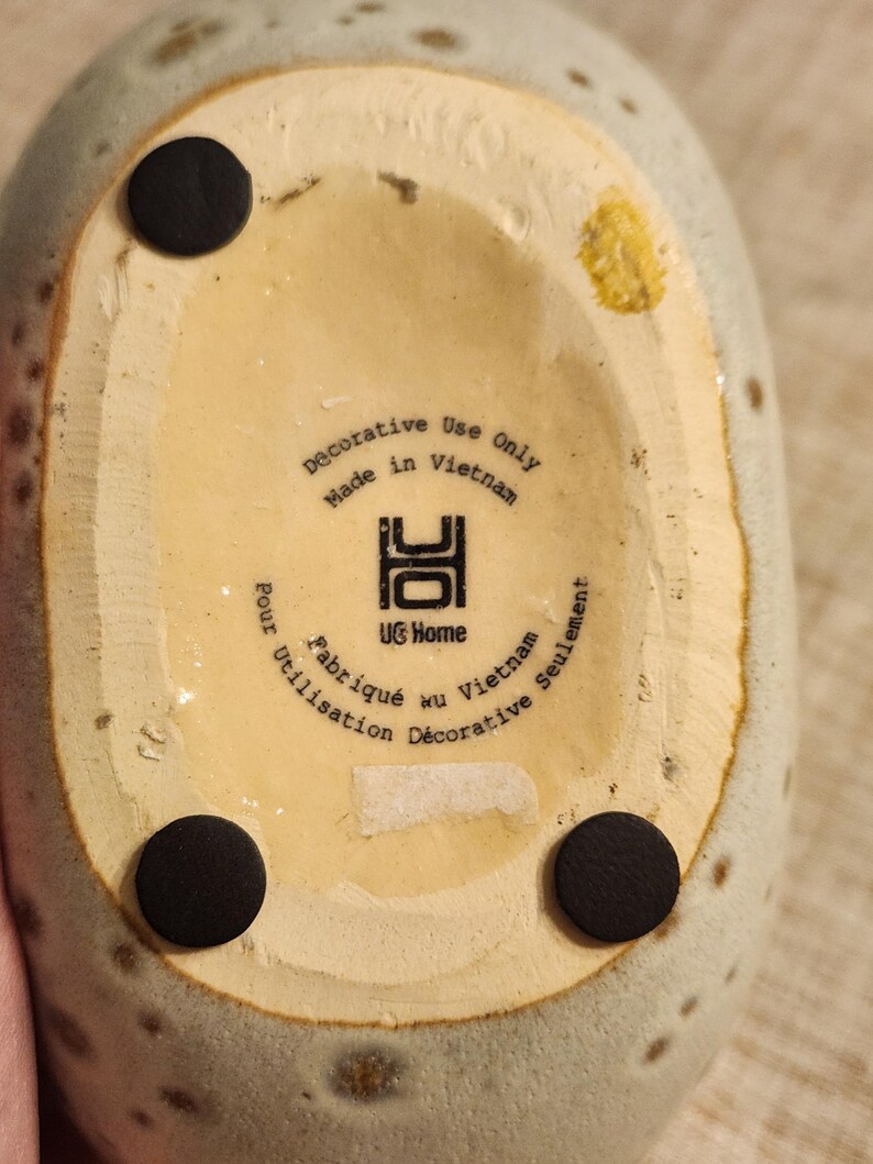 May include: Close-up of the bottom of a speckled, light green ceramic vase. The base is beige with three black circular pads. Text reads "Decorative Use Only Made in Vietnam" and "UG Home".