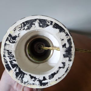 May include: Close-up of the base of a lamp. The lamp base is white with black speckled detailing around the rim. A gold-colored metal fitting is visible inside the base, with a gold electrical cord extending from it.