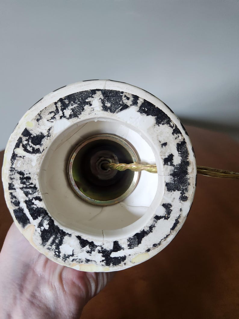 May include: Close-up of the base of a lamp. The lamp base is white with black speckled detailing around the rim. A gold-colored metal fitting is visible inside the base, with a gold electrical cord extending from it.