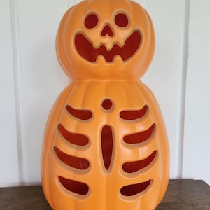 Peut inclure: Une lanterne orange vif en forme de citrouille avec un visage de jack-o'-lantern sculpté sur la partie supérieure et une cage thoracique squelettique découpée sur la partie inférieure. La citrouille a une finition lisse et une tige brune.