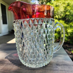 May include: A clear glass pitcher featuring a diamond-cut geometric pattern across the body and a smooth, deep red rim at the top. The vessel includes a curved, transparent glass handle attached to the side, designed for pouring liquids.
