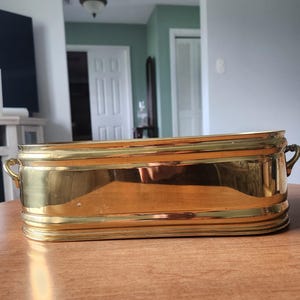 May include: A rectangular brass planter with rounded corners and two small handles. The planter has a polished finish and is approximately 18 inches long.