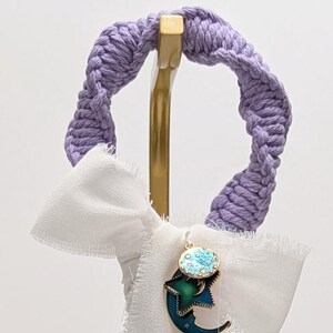 May include: A decorative hanging ornament featuring a lavender macrame top, a white fabric bow, and a charm with a moon and star design. The bottom has tassels in shades of teal, purple, and dark green.