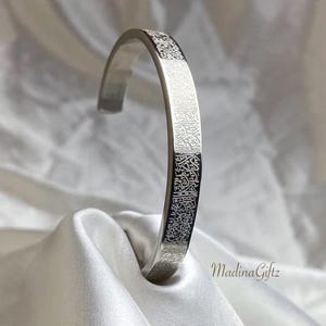 Könnte beinhalten: Ein silbernes Armreif-Armband mit eingravierter arabischer Schrift. Das offene Armband wird auf einer weißen Satin-Oberfläche präsentiert. Die Oberfläche des Armbands ist reflektierend, und die Schrift ist aufwendig detailliert.