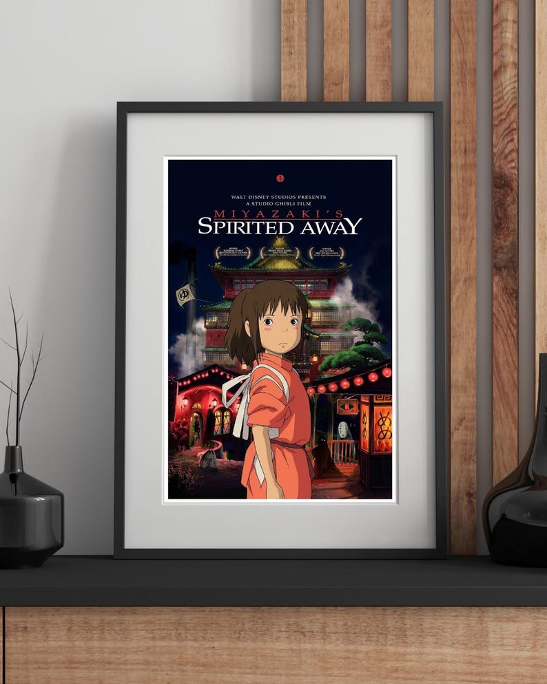 May include: Framed poster of the animated film "Spirited Away." The poster features a young girl in an orange dress standing in front of a detailed building with red lanterns. The film's title and credits are displayed at the top.