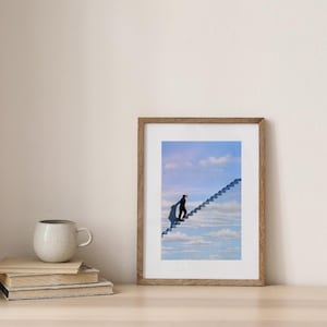May include: A framed art print depicts a surreal scene of a person ascending a staircase that leads into a cloudy sky. The artwork is in a wooden frame and rests on a light-colored wooden surface. A mug and books are in the foreground.