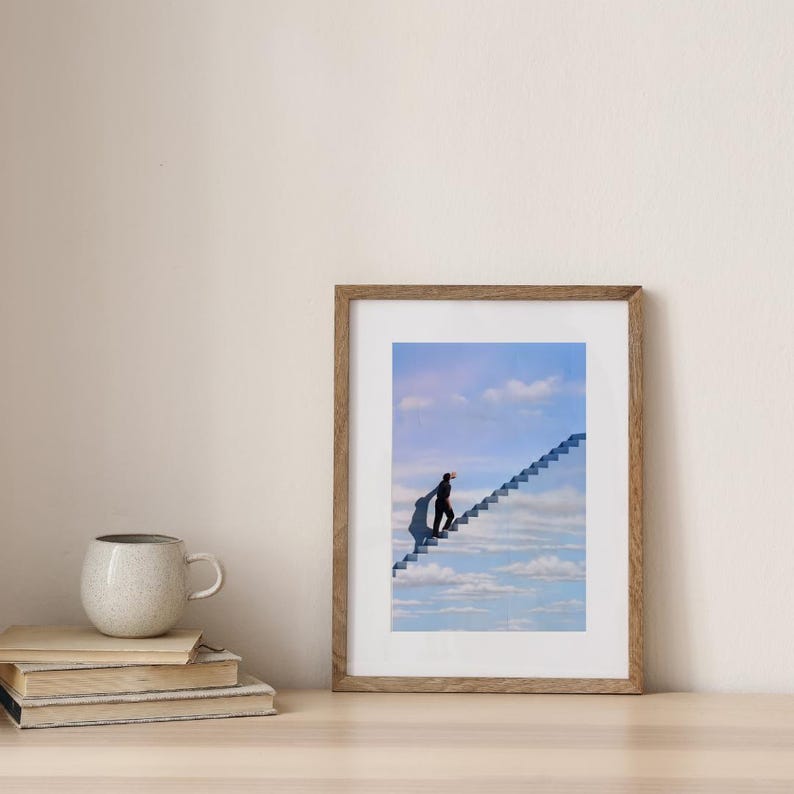 May include: A framed art print depicts a surreal scene of a person ascending a staircase that leads into a cloudy sky. The artwork is in a wooden frame and rests on a light-colored wooden surface. A mug and books are in the foreground.