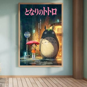 K&ouml;nnte beinhalten: Ein gerahmtes Poster des Zeichentrickfilms "Mein Nachbar Totoro." Das Kunstwerk zeigt ein junges M&auml;dchen mit rotem Regenschirm an einer Bushaltestelle mit Totoro im Regen. Japanischer Text ist oben sichtbar.