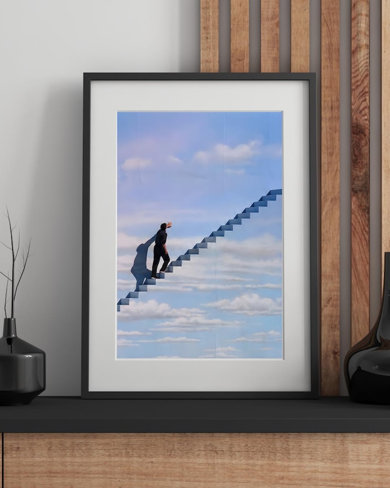 May include: Framed art print featuring a surreal image of a person ascending a staircase that leads into the sky. The artwork is set against a backdrop of a blue sky with fluffy white clouds. The frame is black.