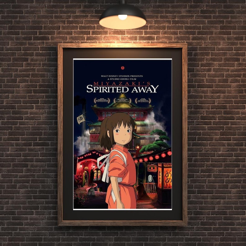 May include: Framed poster of Miyazaki's Spirited Away. The artwork features a young girl in an orange dress standing in front of a traditional Japanese building. The poster is set against a dark brick wall, with a warm light illuminating the scene.