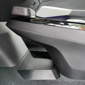 May include: A close-up of a car's interior, showcasing a black, textured armrest with a glossy black trim. A rectangular, dark gray object sits atop the armrest. The design features a sleek, modern aesthetic.