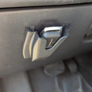 May include: A close-up of a black plastic car component, possibly a latch or clip, with a silver metal piece. The component is mounted on a dark surface, and the design includes curved edges and a recessed area.