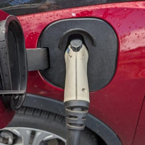 Op de afbeelding: Close-up van een rode elektrische voertuig oplaadpoort met een witte en zwarte oplaadkabel erin. De poort is bedekt met een zwarte klep en de wiel van de auto is zichtbaar op de achtergrond.