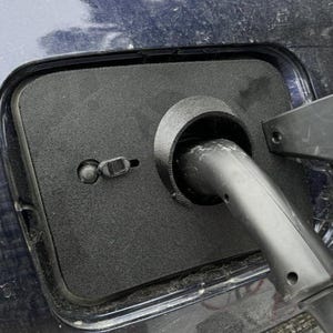 Può includere: Primo piano di una copertura nera della porta di ricarica per veicoli elettrici. La copertura è rettangolare con un'apertura circolare per il cavo di ricarica. Un cavo di ricarica è inserito nella porta. La carrozzeria dell'auto è blu scuro.