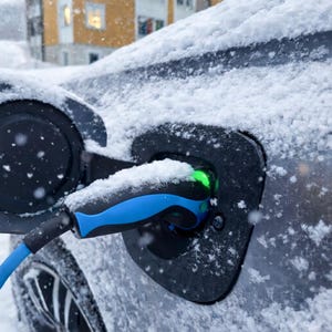 May include: Close-up of a dark grey electric vehicle charging port with a blue and black charging cable plugged in. The port glows green, indicating charging. The car is covered in snow, suggesting winter conditions.