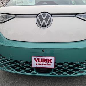 May include: Front view of a white and teal Volkswagen electric vehicle. The VW logo is centered above the teal bumper. A red and white license plate with the text "YURIK accessories" is visible.