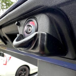 Puede incluir: Primer plano de una cámara de marcha atrás y un asa negros de coche. La lente redonda tiene un reflejo rojo. El asa curvada está integrada en la carrocería. Un coche blanco es visible en el fondo.