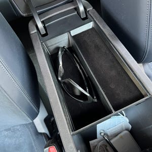 Puede incluir: Consola central de un coche con un compartimento de almacenamiento dividido. El compartimento contiene un par de gafas de sol y una sección forrada de fieltro negro. La consola es negra y gris, con una bisagra plateada.