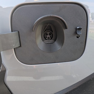 Op de afbeelding: Close-up van een oplaadpoort voor elektrische voertuigen. De poort is verzonken in een grijs paneel aan de buitenkant van het voertuig. De oplaadpoort heeft een cirkelvormig ontwerp met meerdere pinconnectoren. Het paneel heeft een gebogen, metalen handvat.