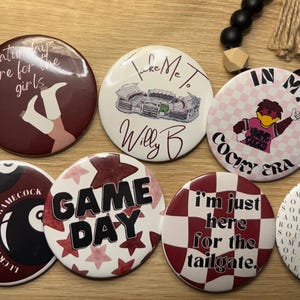Może przedstawiać: Kolekcja okrągłych guzików z różnymi wzorami i tekstami. Guziki zawierają frazy takie jak "BEAT OKLAHOMA", "GAME DAY" i "I'm just here for the tailgate". Kolory to między innymi bordowy, biały i czarny.