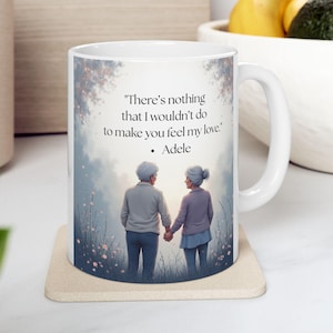 May include: White ceramic mug with a wraparound design featuring an elderly couple holding hands, with the quote "There's nothing that I wouldn't do to make you feel my love." by Adele. The mug sits on a beige coaster.