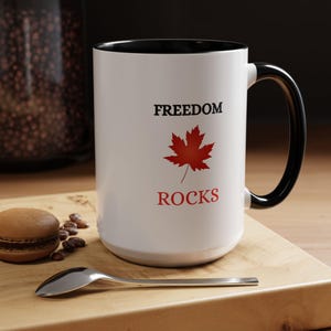 Puede incluir: Taza de cerámica blanca con interior y asa negros. La taza presenta las palabras "FREEDOM ROCKS" en negro y rojo, con un gráfico de hoja de arce roja. La taza está sobre una superficie de madera con una cuchara, granos de café y un pastel.